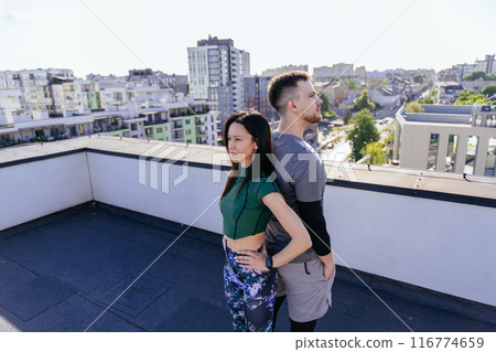 Athletic man and flexible woman standing back to back in morning in rooftop on urban background. Athletic man and flexible woman standing back to back in morning in rooftop on urban background. 116774659
