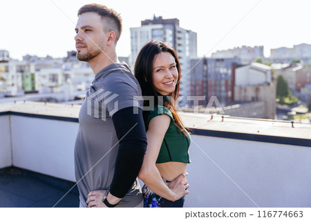 Lovely couple dressed in athletic wear, are standing back-to-back on a rooftop urban background. They both are looking over their shoulders Lovely couple dressed in athletic wear, are standing back-to-back on a rooftop urban background. They both are looking over their shoulders 116774663