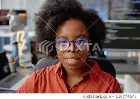 Close up portrait of curly female African American software developer wearing glasses smiling while looking at camera in computer programmer office Close up portrait of curly female African American software developer wearing glasses smiling while looking at camera in computer programmer office 116774675