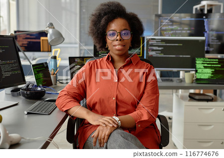 Medium portrait shot of curvy female African American VR engineer in glasses smiling looking at camera while sitting at working desk with multiple computer screens in office, copy space Medium portrait shot of curvy female African American VR engineer in glasses smiling looking at camera while sitting at working desk with multiple computer screens in office, copy space 116774676