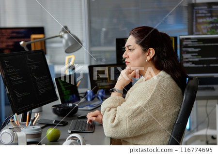 Side view shot of thoughtful female Middle Eastern reviewing code fixing errors while sitting at workplace with digital devices and green apple at desk in IT company office, copy space Side view shot of thoughtful female Middle Eastern reviewing code fixing errors while sitting at workplace with digital devices and green apple at desk in IT company office, copy space 116774695