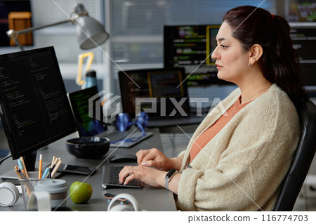 Side view shot of female Middle Eastern software engineer writing code on computer with multiple screens while sitting at desk with green apple at hand in IT company office, copy space 116774703