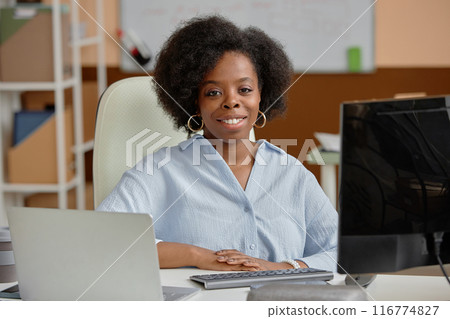 Portrait shot of successful smiling Black businesswoman looking at camera while sitting at working desk with computers in coworking office 116774827