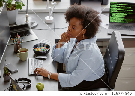 High-angle shot of thoughtful female African American senior IT specialist reading computer screen, while sitting at working desk with multiple monitors and candy bowl at hand in office 116774840