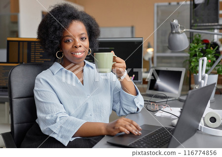 Portrait shot of smiling young Black woman working in IT development office sitting at work desk while having cup of coffee looking at camera, copy space Portrait shot of smiling young Black woman working in IT development office sitting at work desk while having cup of coffee looking at camera, copy space 116774856