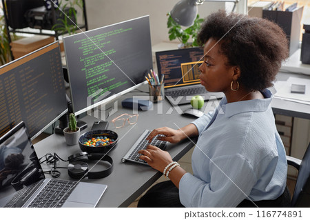 High-angle side view shot of African American female programmer working on computer reading multiple screens with code lines running 116774891