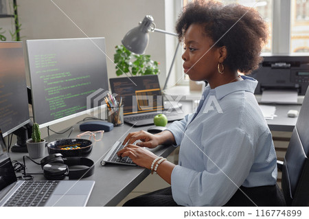 Side view of young African American female software developer using computer writing code while sitting at desk with multiple screens in office 116774899
