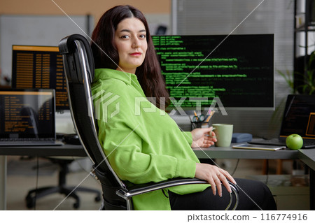 Portrait of calm higher-weight female Middle Eastern programmer in shirt of green color looking at camera, while sitting in chair at desk with multiple computer screens in office Portrait of calm higher-weight female Middle Eastern programmer in shirt of green color looking at camera, while sitting in chair at desk with multiple computer screens in office 116774916