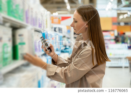 A woman in a beige shirt stands in the aisle of a grocery store, reaching for a pack of adult diapers. There are a lot of goods in the aisle, and the shelves are filled with a variety of goods. A woman in a beige shirt stands in the aisle of a grocery store, reaching for a pack of adult diapers. There are a lot of goods in the aisle, and the shelves are filled with a variety of goods. 116776065