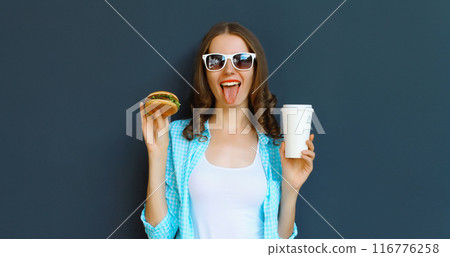 Happy cheerful young woman eating burger fast food holding cup of coffee, teenage girl having fun 116776258