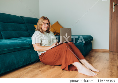 Nice beautiful woman in glasses working at the notebook near the sofa at home. Concepts of education, work and comfort. 30s female working indoors. Full body length 116776405
