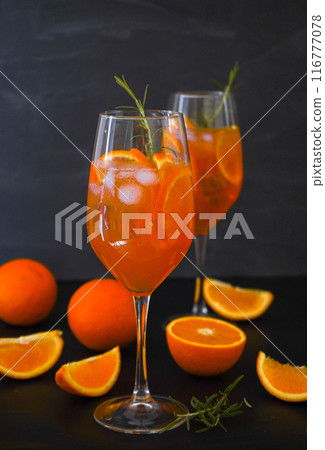 Aperol spritz is a summer drink served in a glass with aperol, prosecco, soda and orange slice. Glasses of icy cocktail in a wine glass and a rosemary branch at the bar. Black background. Refreshing Aperol spritz is a summer drink served in a glass with aperol, prosecco, soda and orange slice. Glasses of icy cocktail in a wine glass and a rosemary branch at the bar. Black background. Refreshing 116777078