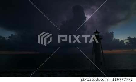 White tall lighthouse on sea shore with blinking light at stormy night for commercial vessels navigation. Thunderstorm with lightnings over ocean water posing danger for ships White tall lighthouse on sea shore with blinking light at stormy night for commercial vessels navigation. Thunderstorm with lightnings over ocean water posing danger for ships 116777772