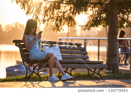 Young woman sitting on park bench browsing her cellphone outdoors on warm summer evening. Communication and mobile connection concept. 116778483