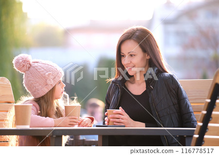 Young mother and her child daughter having good time together sitting at street cafe with hot drinks on sunny fall day. Happiness in family relations concept 116778517