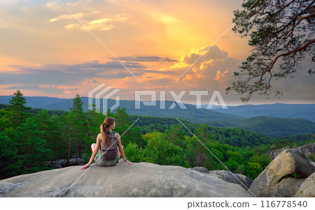 Woman hiker seated alone on rocky mountain cliff enjoying view of evening nature on wilderness trail. Active way of life concept Woman hiker seated alone on rocky mountain cliff enjoying view of evening nature on wilderness trail. Active way of life concept 116778540
