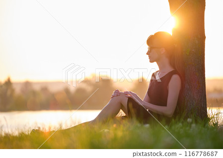 Lonely woman sitting alone on green grass lawn leaning to tree trunk on lake shore on warm evening. Solitude and relaxing in nature concept 116778687