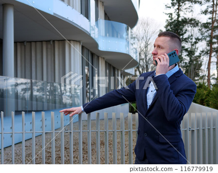 A Businessman engages in phone conversation outside an office building, dressed in formal business attire. The Businessman's professional demeanor is evident as he discusses important matters. A Businessman engages in phone conversation outside an office building, dressed in formal business attire. The Businessman's professional demeanor is evident as he discusses important matters. 116779919