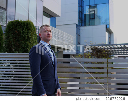 A Businessman in business attire stands thoughtfully with an office building backdrop. The Businessman's pose reflects his strategic thinking. This scene captures the Businessman's professionalism 116779926
