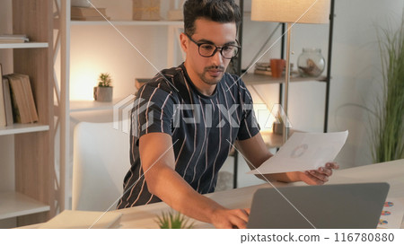 Distance job. Work documentation. Busy focused business man studying paper report data keeping record on laptop sitting at desk in home office. 116780880