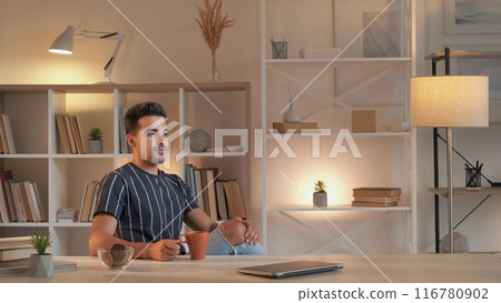 Work rest. Coffee break. Enjoying leisure. Relaxed man drinking warm beverage from cup sitting at desk with laptop in modern home interior copy space. 116780902