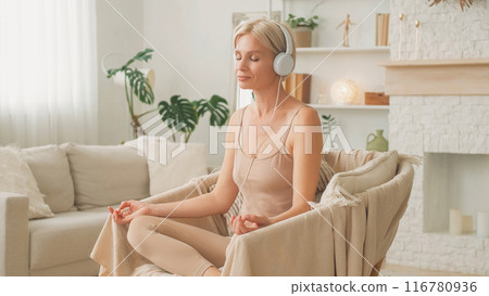 Audio meditation. Peaceful balance. Wellness leisure. Relaxed woman in headphones sitting in comfortable position in armchair listening to mantra at home. 116780936