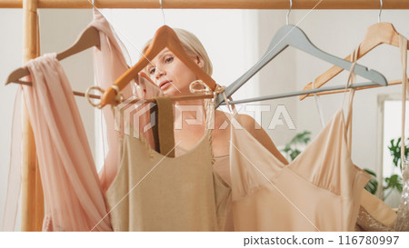Elegant look. Clothes shopping. Beautiful customer woman choosing trendy beige dress on hanger for holiday party in light interior showroom. Elegant look. Clothes shopping. Beautiful customer woman choosing trendy beige dress on hanger for holiday party in light interior showroom. 116780997