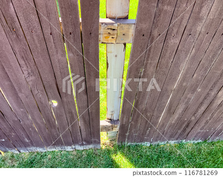 A backyard wooden fence showing a small gap between its panels. The brown fence, weathered with time, is highlighted by the sunlit grass visible through the gap. This image captures the rustic charm 116781269