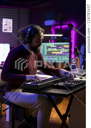 Songwriter plays midi controller piano notes in his home studio, using daw software on computer to record audio files. Young musician playing tunes on electronic keyboard, post production. 116781437