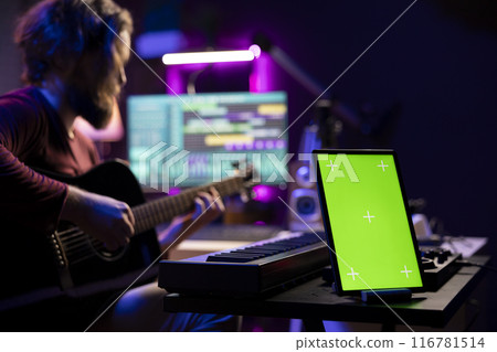 Audio engineer playing guitar and recording tunes on daw software, skilled artist learning to play new songs and strings accords. Music producer uses greenscreen on tablet, home studio. 116781514
