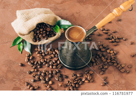 A bag of coffee beans is poured onto table, cup of freshly prepared coffee, brown background 116781977