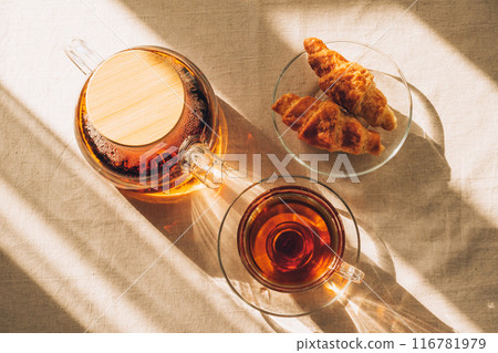 Top view of glass teapot with brewed tea, cup of tea and croissants on table under sunlight casting shadows Top view of glass teapot with brewed tea, cup of tea and croissants on table under sunlight casting shadows 116781979