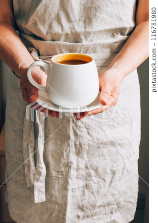 A man in linen apron holding white cup of coffee on saucer. Hot drink in hands, close-up 116781980