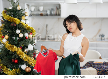 Sad young Asian girl trying to choose a dress standing next to Xmas tree 116783314