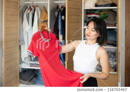 Happy young Asian girl trying to choose a dress standing by wardrobe Happy young Asian girl trying to choose a dress standing by wardrobe 116783467