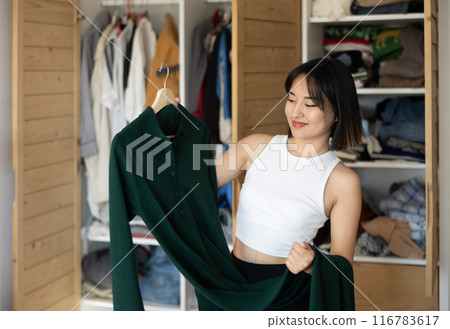 Happy young Asian girl trying to choose a dress standing by wardrobe 116783617