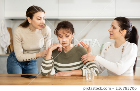 Sad woman sits silently in kitchen, adult girls sympathize and comfort her Sad woman sits silently in kitchen, adult girls sympathize and comfort her 116783706
