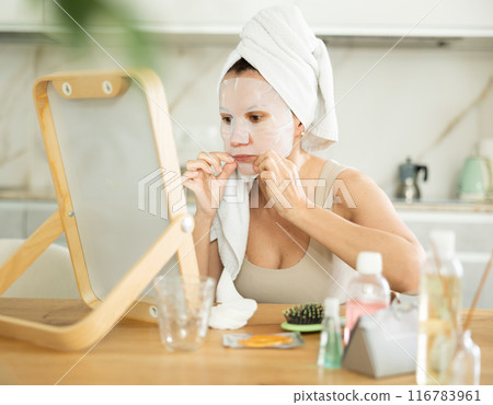 Middle-aged woman applying tissue mask on her facial skin sitting in front of the mirror Middle-aged woman applying tissue mask on her facial skin sitting in front of the mirror 116783961