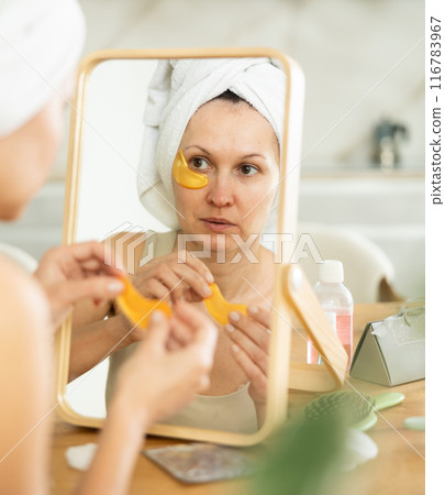 Woman in towel turban applying golden collagen eye patches 116783967
