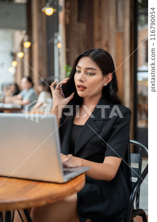 A confident Asian businesswoman is talking on the phone while working on her laptop at a cafe. A confident Asian businesswoman is talking on the phone while working on her laptop at a cafe. 116790054