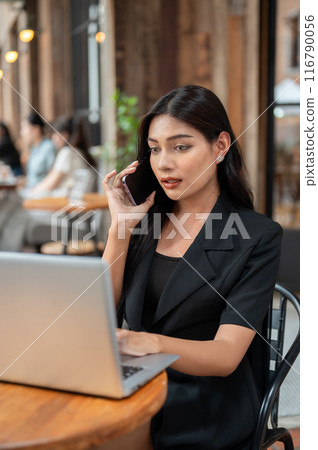 A confident Asian businesswoman is talking on the phone while working on her laptop at a cafe. 116790056