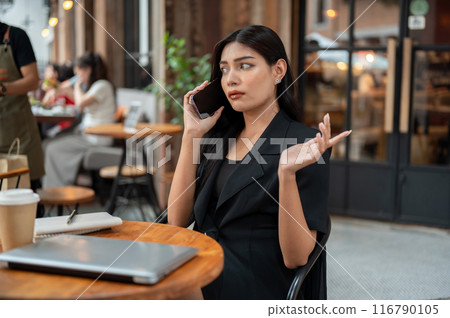 Uncertain, thoughtful Asian businesswoman is talking on the phone while sitting at a table of a cafe 116790105