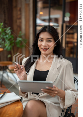 A gorgeous Asian businesswoman is sitting at an outdoor table of a cafe, holding a digital tablet. A gorgeous Asian businesswoman is sitting at an outdoor table of a cafe, holding a digital tablet. 116790143