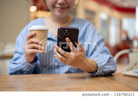 A cropped image of an Asian woman using her smartphone while sitting at a table in a coffee shop. A cropped image of an Asian woman using her smartphone while sitting at a table in a coffee shop. 116790221
