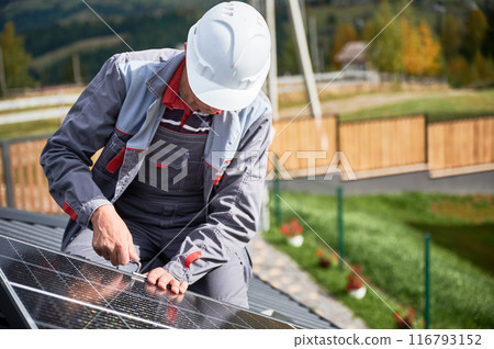 Man technician mounting photovoltaic solar panels on roof of house. Mounter in helmet installing solar module system with help of hex key. Concept of alternative, renewable energy. 116793152