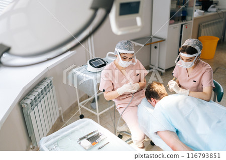 Top view of two female doctors extracting hair follicles from donor area of male patient lying on stomach for hair transplantation in clinic. Process of hair transplant with FUE Sapphire method. 116793851
