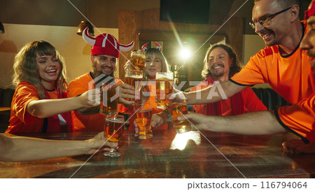 Friends, football fans in orange shirts raise their glasses in celebratory toast, their faces beaming with happiness and energy. 116794064