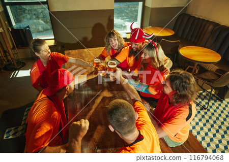 Fans in matching orange shirts and fun hats cheerfully toast with their beers at bar, celebrating with wide smiles. 116794068