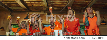 Banner. At a bar, excited people in orange shirts celebrate with raised glasses victory of favorite sport team while watching its match on TV. 116794096