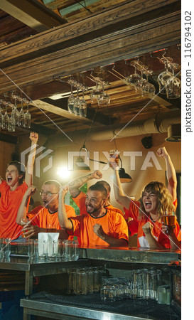 Excited young men and women in orange shirts celebrate at bar, raising their beers and cheering, victory of favorite sport team. Excited young men and women in orange shirts celebrate at bar, raising their beers and cheering, victory of favorite sport team. 116794102